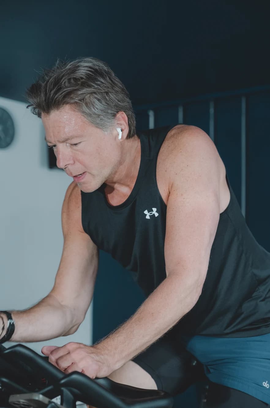 A man wearing a black sleeveless top pedals an exercise bike in a dimly lit gym.