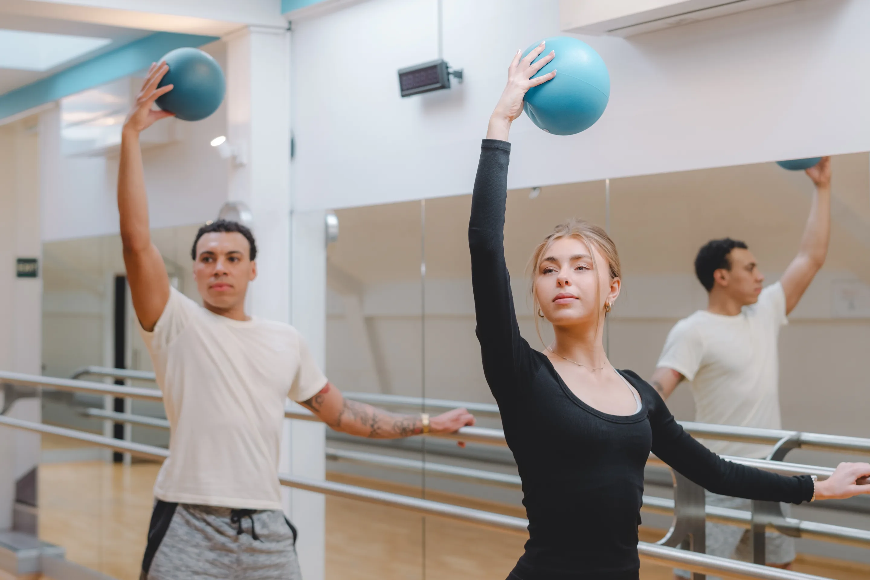 Two dancers in a mirrored studio raise blue exercise balls overhead while extending their free arms.