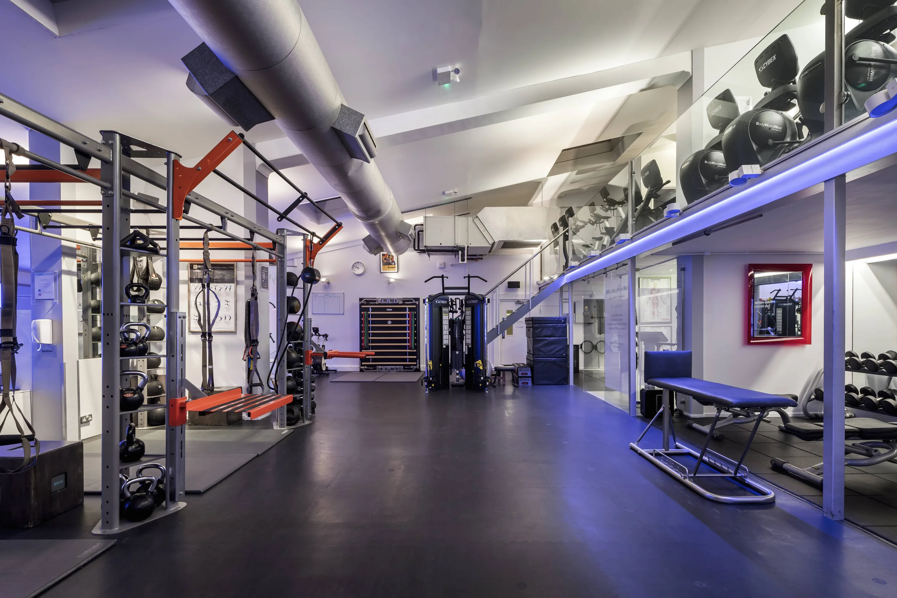 A modern gym interior with weight racks, cable machines and a padded bench under blue accent lighting.