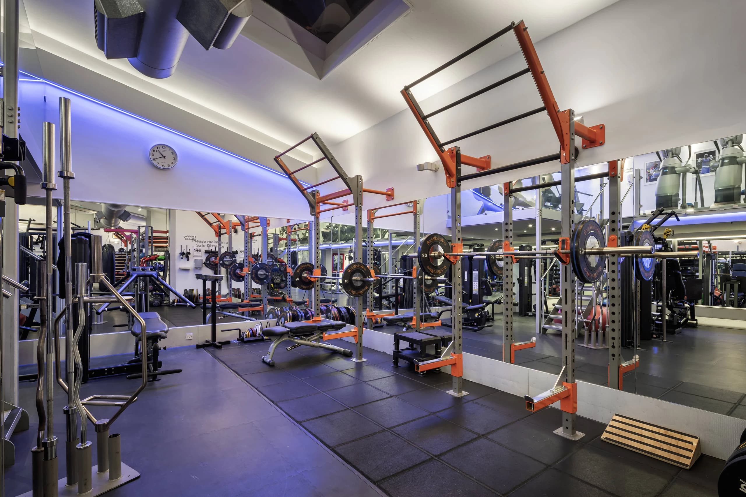 A brightly lit gym with weight racks, barbells and benches reflected in mirrored walls, beneath blue accent lighting.