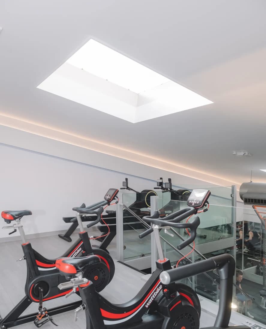 Modern gym with black and red exercise bikes and a treadmill behind a glass railing beneath a bright skylight.
