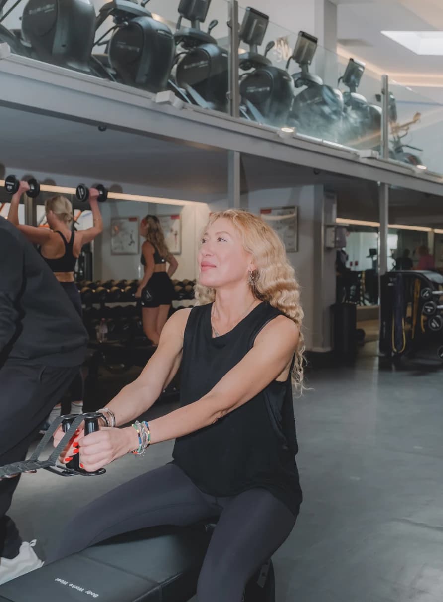 A woman uses a seated rowing machine in a gym, with other people lifting weights behind her.