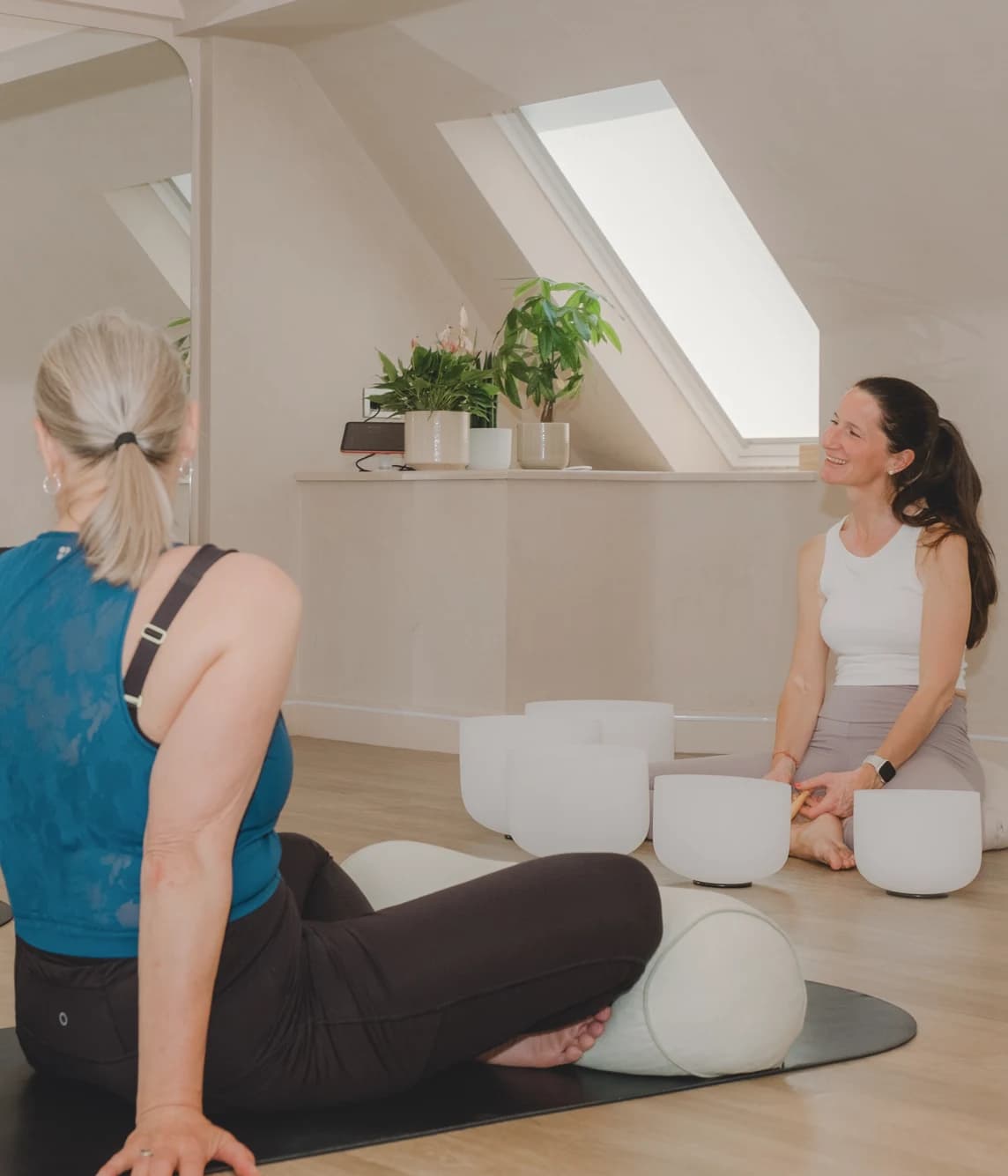 Two women exercise on yoga mats in a bright attic studio with a skylight, plants and white fitness equipment.