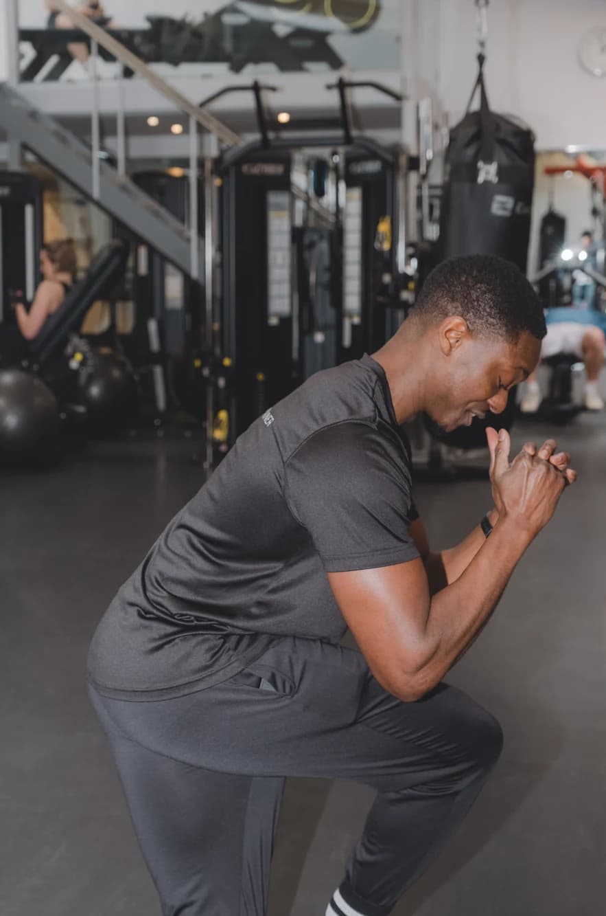 A man in grey sportswear performs a kneeling lunge in a gym, with weight machines and other people in the background.